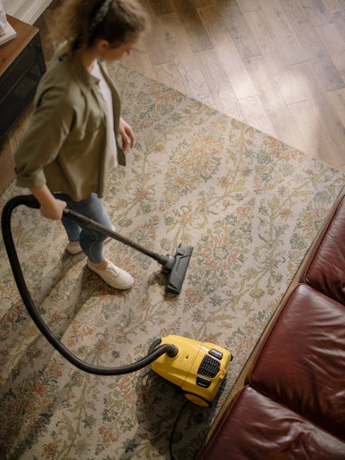 A woman using a yellow vacuum cleaner with a black hose to deep clean an ornate patterned rug on a wooden floor in a living room. She is wearing casual clothing including a beige jacket, jeans, and wh