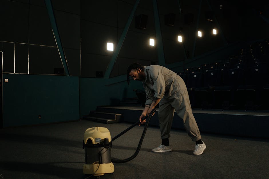 A woman using a yellow vacuum cleaner with a black hose to deep clean an ornate patterned rug on a wooden floor in a living room. She is wearing casual clothing including a beige jacket, jeans, and wh