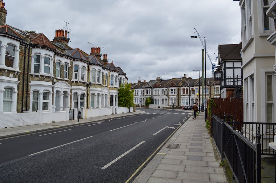A residential street scene near Grahame Park and Burnt Oak featuring a row of Victorian-style terraced houses with white painted facades, large bay windows, and red-tiled roofs. The pavement on the ri